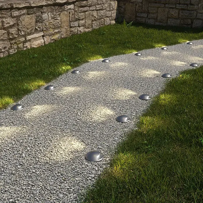 Lucande LED-Gartenstrahler Helene - 3.000 K, 8,8 x 8,8 x 9,5 cm - Silberne Dom-Wegleuchten beleuchten einen Kiesweg neben Gras und einer Steinmauer.