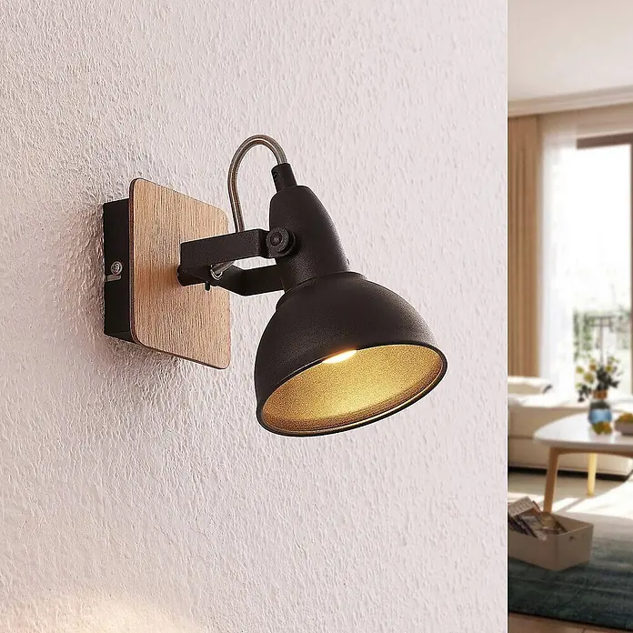 Lindby Wandstrahler Aylis - Holz dunkel, schwarz, E14 - Schwarze Metall-Wandleuchte mit Holzsockel und verstellbarem Kopf, beleuchtet eine strukturierte Wand in einem modernen Wohnraum.