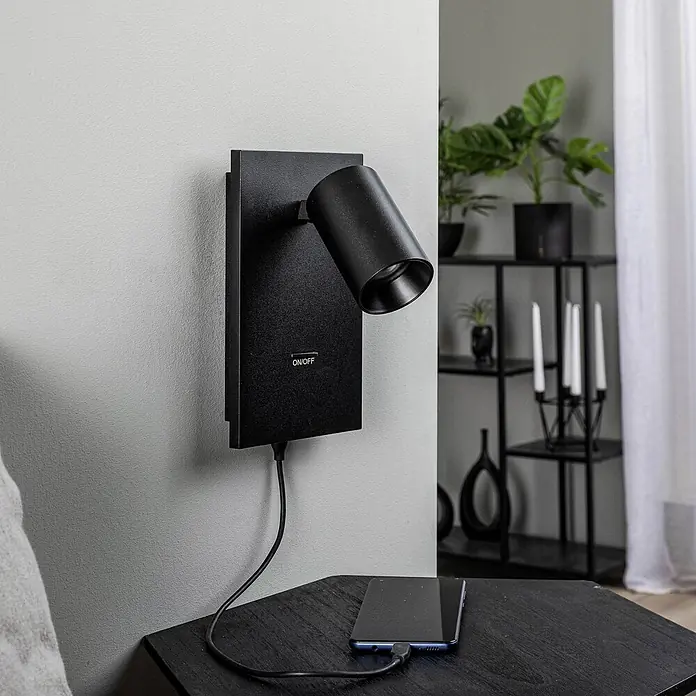 Arcchio Wandstrahler Brinja - schwarz, schwarz, GU10 - Schwarze wandmontierte Leselampe mit verstellbarem Kopf und ON/OFF-Schalter, lädt ein Smartphone auf einem dunklen Holznachttisch.
