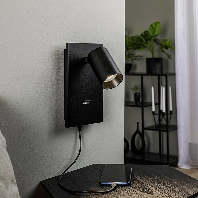 Arcchio Wandstrahler Brinja - schwarz, schwarz, GU10 - Schwarze Wandleuchte mit integriertem Telefonladegerät, beleuchtet einen Nachttisch, auf dem ein Smartphone lädt.