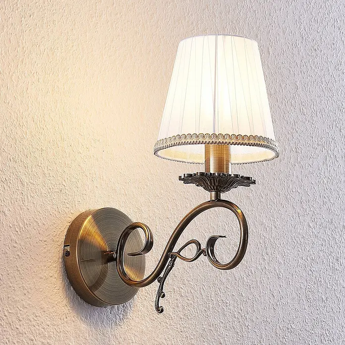 Lindby Wandleuchte Finnick - bronze / altmessing, weiß / opal, E14 - Beleuchtete Wandleuchte aus Antikbronze mit weißem, plissiertem Stoffschirm an einer strukturierten Wand.