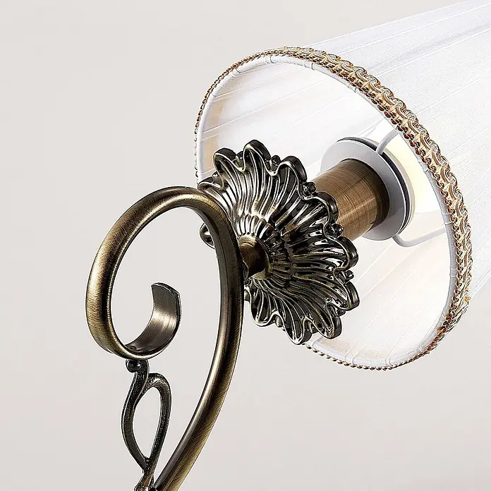 Lindby Wandleuchte Finnick - bronze / altmessing, weiß / opal, E14 - Nahaufnahme einer Lampe mit antikisiertem Messing-Rankenarm, floralem Fuß und weißem Plissee-Stoffschirm mit Goldbesatz.