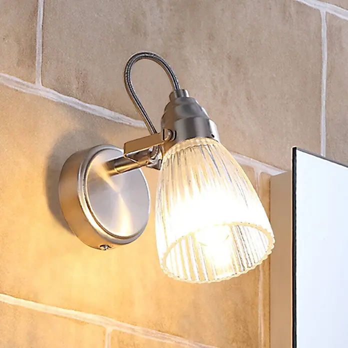 Lindby Wandleuchte Kara - L x B x H: 14 x 9 x 21 cm, alu / grau / zink, G9 - Wandleuchte mit gebürstetem Metallsockel und geriffelter Glasschirm, strahlt warmes Licht an einer gefliesten Wand aus.