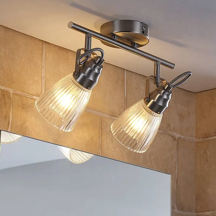 Lindby Wandleuchte Kara - alu / grau / zink, G9 - Gebürstete Nickeldeckenleuchte mit zwei gerippten Glasschirmen, beleuchtet eine geflieste Wand über einem Spiegel.