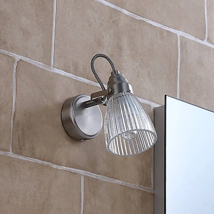 Lindby Wandleuchte Kara - L x B x H: 14 x 9 x 21 cm, alu / grau / zink, G9 - Wandleuchte mit gebürstetem Metallfuß und geripptem Glasschirm, an einer Fliesenwand neben einem Spiegel montiert.
