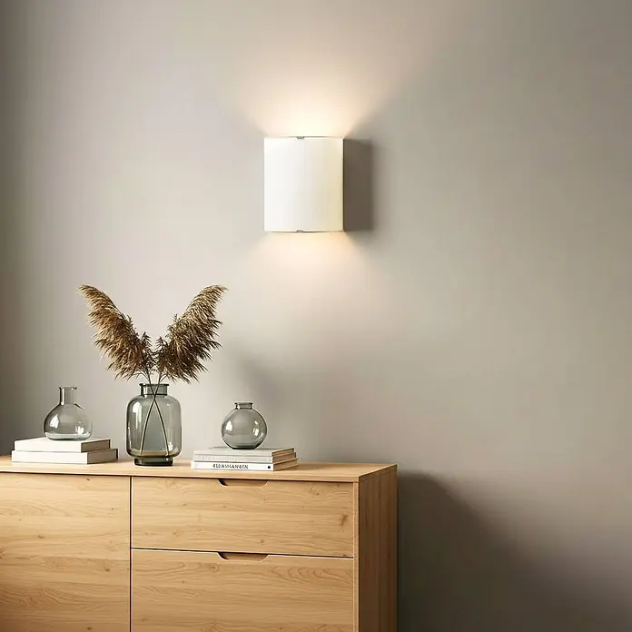 Lindby Wandleuchte Phil - weiß / opal, E27 - Weiße zylindrische Wandleuchte, die an einer Wand über einer Holzkommode Licht nach oben und unten wirft.