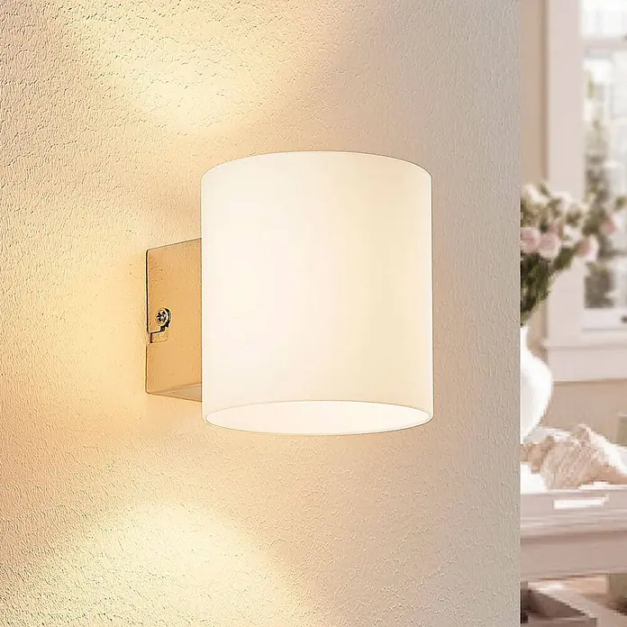 Lindby Wandleuchte Gerrit - L x B x H: 12,2 x 9,2 x 9,5 cm, weiß / opal, G9 - Wandleuchte mit weißem zylindrischem Stoffschirm und goldfarbenem Metallsockel, die eine strukturierte Wand beleuchtet.