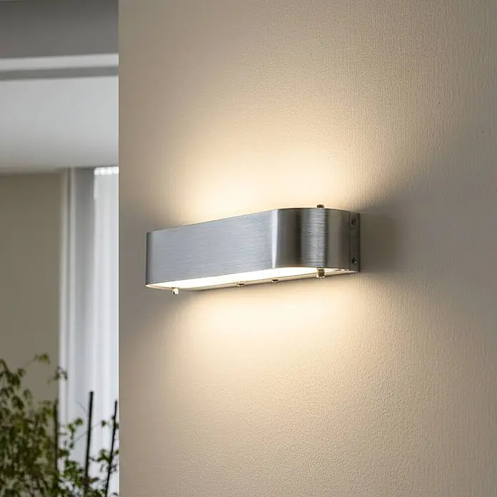 Lindby Wandleuchte Nika - alu / grau / zink, alu / grau / zink, E14 - Gebürstete Aluminium-Wandleuchte mit abgerundeten Enden, die warmes Licht auf eine strukturierte Wand wirft.