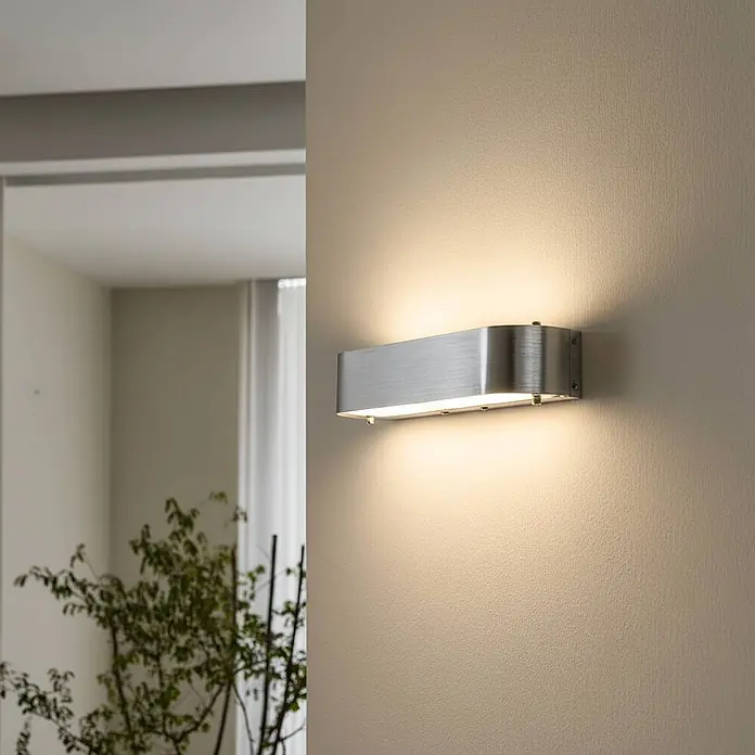 Lindby Wandleuchte Nika - alu / grau / zink, alu / grau / zink, E14 - Gebürstete Metall-Wandleuchte strahlt warmes Licht auf eine strukturierte beigefarbene Wand.