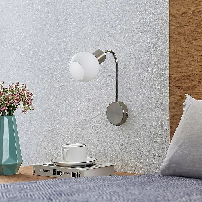 Lindby Wandleuchte Lioma - alu / grau / zink, E14 - Wandleseleuchte aus gebürstetem Nickel mit flexiblem Schwanenhals und weißer Glaskugel, neben einem Bett platziert.