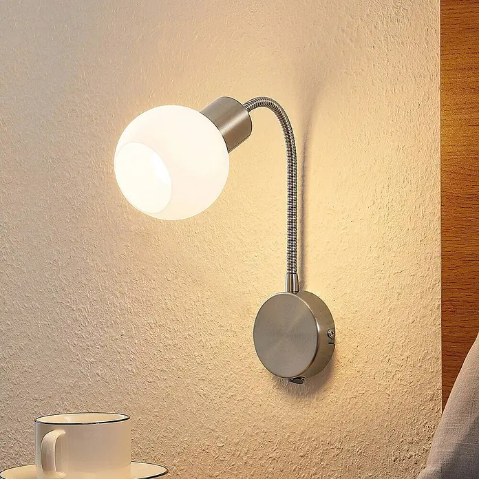 Lindby Wandleuchte Lioma - alu / grau / zink, E14 - Wandleseleuchte mit gebürsteter Nickelbasis, flexiblem Schwanenhals und Milchglas-Kugelschirm, die einen Nachttisch beleuchtet.