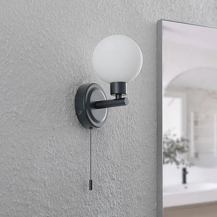 Lindby Wandleuchte Zaniba - L x B x H: 12,4 x 10 x 17,7 cm, schwarz, G9 - Dunkelgraue Wandleuchte mit kugelförmigem Milchglasschirm und Zugschalter, an einer strukturierten Wand neben einem Spiegel montiert.