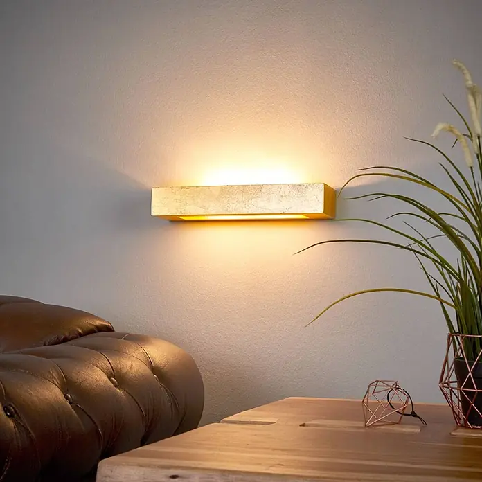 Lindby Wandleuchte Emina - L x B x H: 9,5 x 36 x 6 cm, messing / gold, E14 - Goldene rechteckige Wandleuchte, die eine Wand über einem Sofa und neben einer Pflanze beleuchtet.
