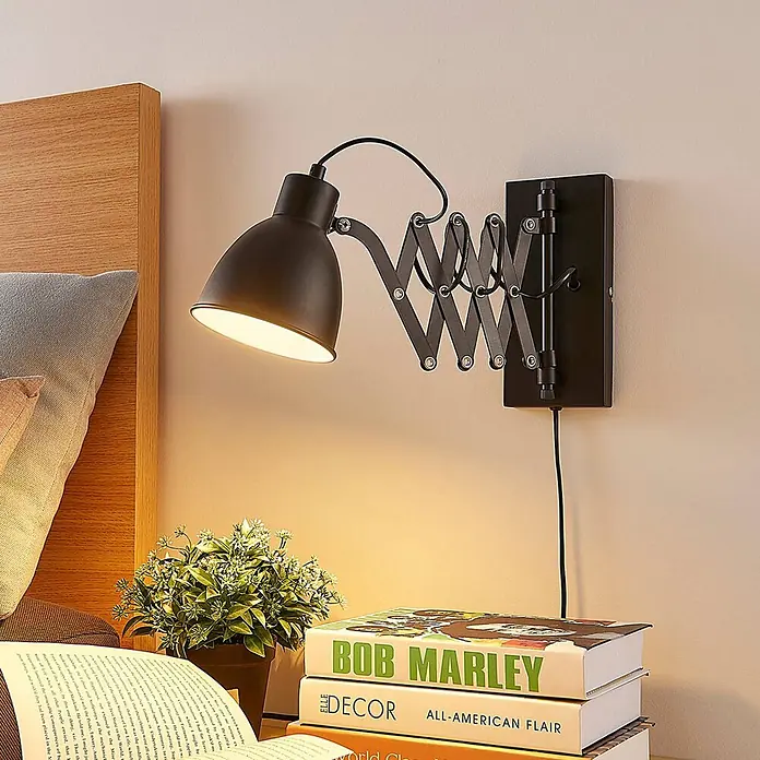 Lindby Wandleuchte Merle - schwarz, schwarz, E14 - Schwarze Wandlampe aus Metall mit Scherenarm und konischem Schirm beleuchtet einen Nachttisch neben einem Holzbett.