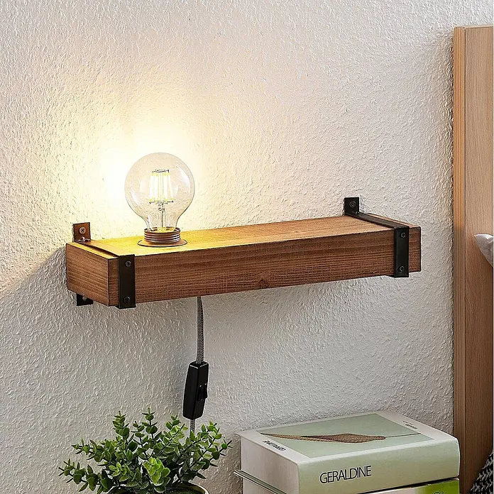 Lindby Wandleuchte Sverina - L x B x H: 12 x 40 x 10 cm, Holz dunkel, E27 - Ein hölzernes Wandregal mit klassischer Glühbirne und schwarzen Metallhalterungen, als Nachttischlampe über Büchern und einer Pflanze.