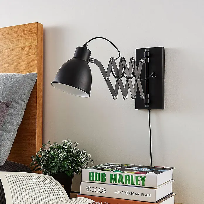 Lindby Wandleuchte Merle - schwarz, schwarz, E14 - Schwarze Wandleuchte mit Scherengelenk neben einem Bett, die Bücher auf einem Nachttisch beleuchtet.