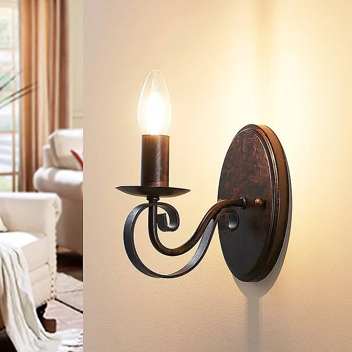 Lucande Wandleuchte Caleb - braun / rost, braun / rost, E14 - Eine vintage-inspirierte Wandleuchte aus dunkler Bronze mit eleganter Schnörkelarbeit und einer kerzenförmigen Glühbirne an einer hellen Wand.