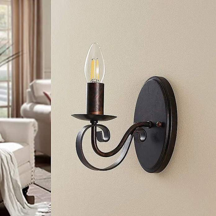 Lucande Wandleuchte Caleb - braun / rost, braun / rost, E14 - Eine antike bronzefarbene Wandleuchte mit LED-Kerzenbirne an cremefarbener Wand, die ein Wohnzimmer aufwertet.