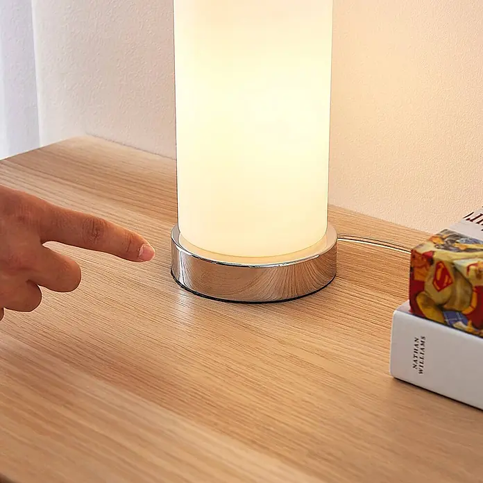 Lindby Tischleuchte Elianna - weiß / opal, E14 - Ein Finger berührt den Chromsockel einer leuchtenden zylindrischen Tischlampe auf einer Holzoberfläche.