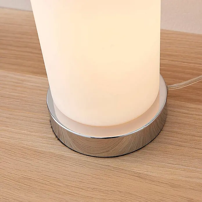 Lindby Tischleuchte Elianna - weiß / opal, E14 - Nahaufnahme einer Lampe mit leuchtend weißem Zylinderschirm und glänzendem Chromfuß auf einem Holztisch, mit sichtbarem Kabel.