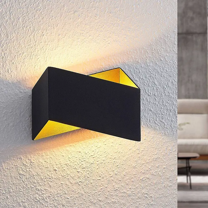 Arcchio Wandleuchte Assona - schwarz, G9 - Moderne schwarz-goldene Wandleuchte beleuchtet eine strukturierte Wand mit warmem Up- und Downlight.
