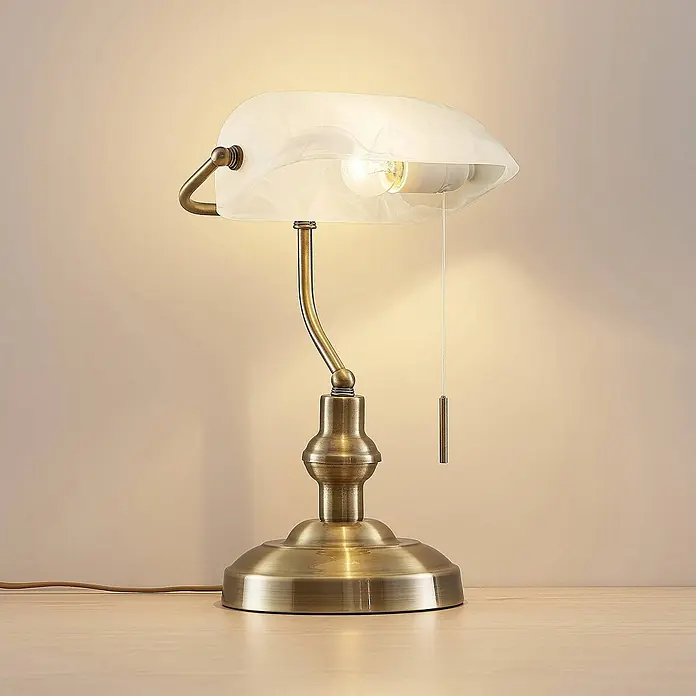 Lindby Tischleuchte Profina - L x B x H: 18,5 x 27,2 x 38,5 cm, bronze / altmessing, weiß / opal, E27 - Eine antike Messing-Bankerlampe mit mattweißem Glasschirm und Zugschalter auf einem Holzschreibtisch.