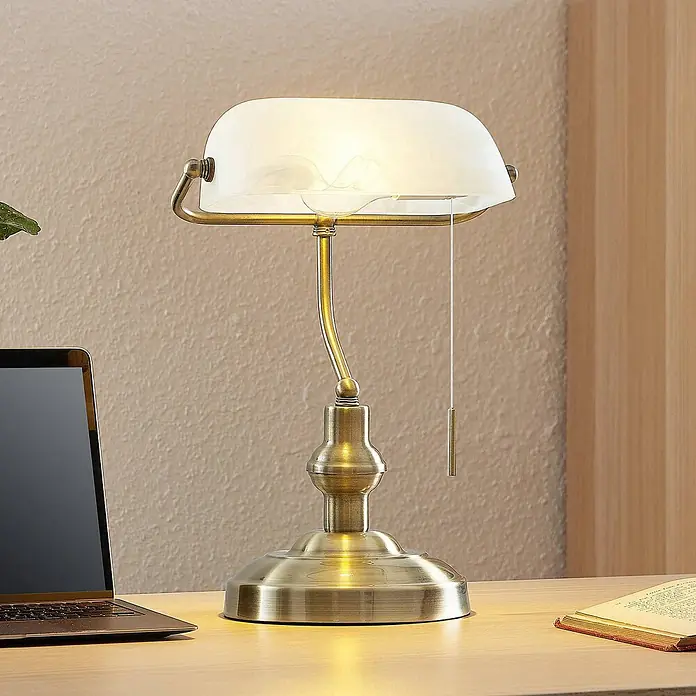 Lindby Tischleuchte Profina - L x B x H: 18,5 x 27,2 x 38,5 cm, bronze / altmessing, weiß / opal, E27 - Antike Messing-Bankerlampe mit weißem Glasschirm, beleuchtet einen Holzschreibtisch mit Laptop und Buch.