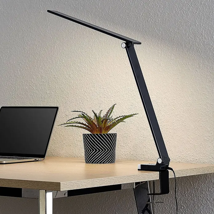 Prios Tischleuchte Tamarin - schwarz - Schwarze LED-Schreibtischlampe mit verstellbarem Arm und Klemmfuß, leuchtet einen Holzschreibtisch mit Laptop aus.