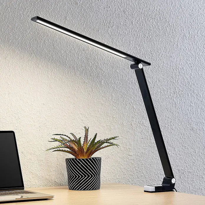 Prios Tischleuchte Tamarin - schwarz - Verstellbare schwarze LED-Schreibtischlampe beleuchtet einen Holztisch neben Laptop und Pflanze.