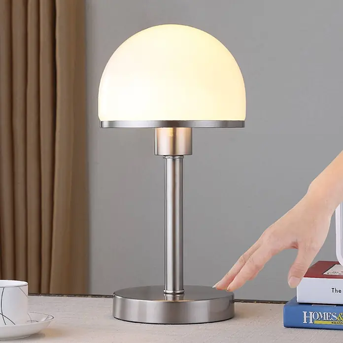Lindby Tischleuchte Jolie - alu / grau / zink, E27 - Silberne Tischlampe aus gebürstetem Metall mit Milchglasschirm; eine Hand berührt die Basis.