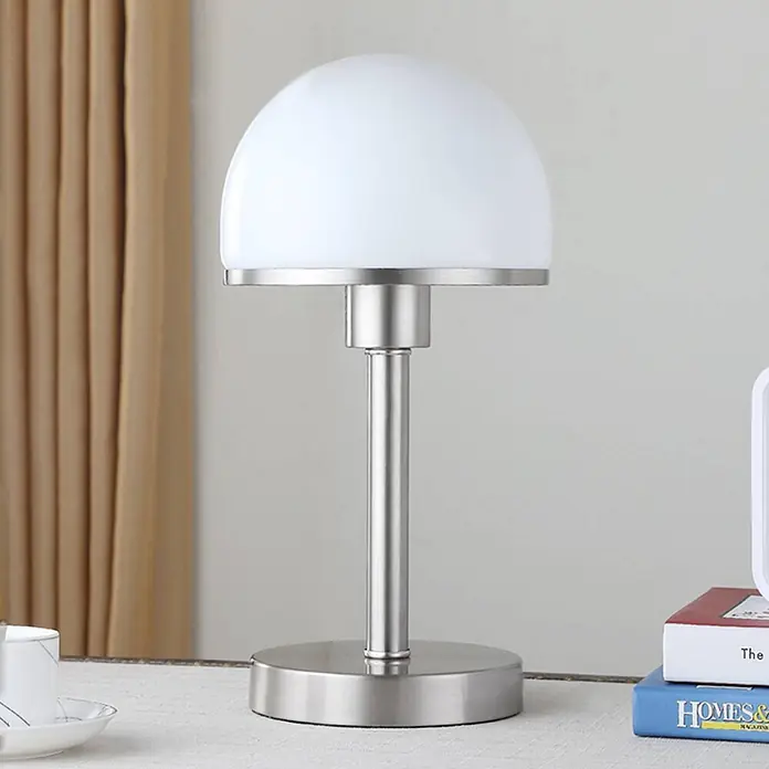 Lindby Tischleuchte Jolie - alu / grau / zink, E27 - Moderne Tischlampe mit gebürstetem Metallsockel und Stiel, mit einem halbkugelförmigen weißen Glasschirm, auf einem Tisch platziert.