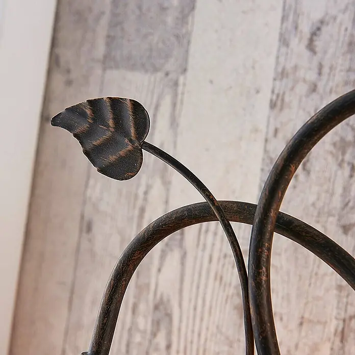 Lindby Tischleuchte Stefania - L x B x H: 28 x 28 x 40 cm, braun / rost, E14 - Nahaufnahme eines dekorativen Metallornaments mit gealtertem dunklem Finish und Bronzeakzenten, geformt wie ein Blatt und gebogene Äste, vor hellem Holzgrund.