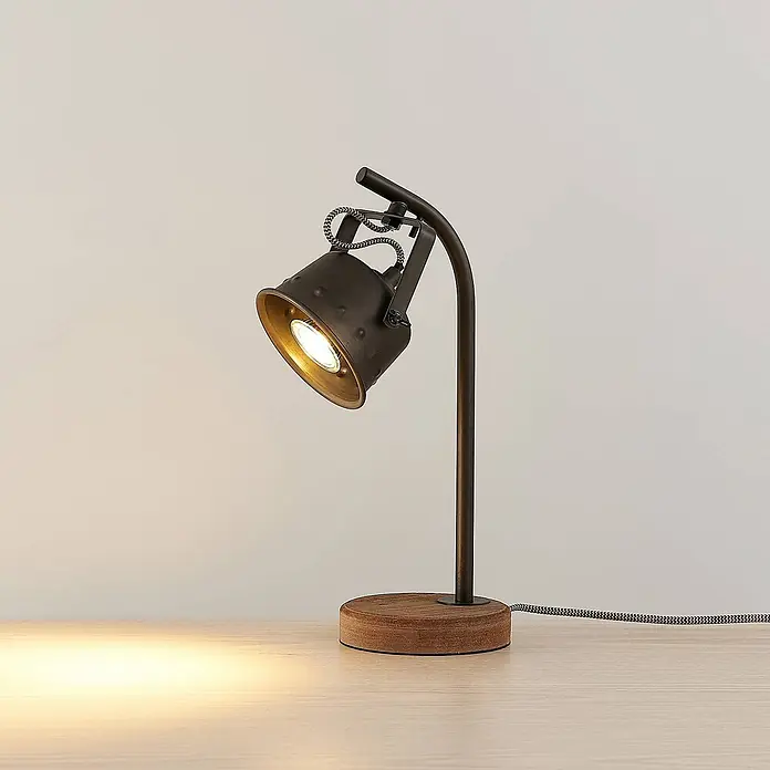 Lindby Tischleuchte Rubinjo - L x B x H: 20 x 13 x 36,5 cm, Holz dunkel, schwarz, GU10 - Dunkle Metall-Tischleuchte mit rundem Holzsockel und geflochtenem Kabel beleuchtet eine helle Holzfläche.