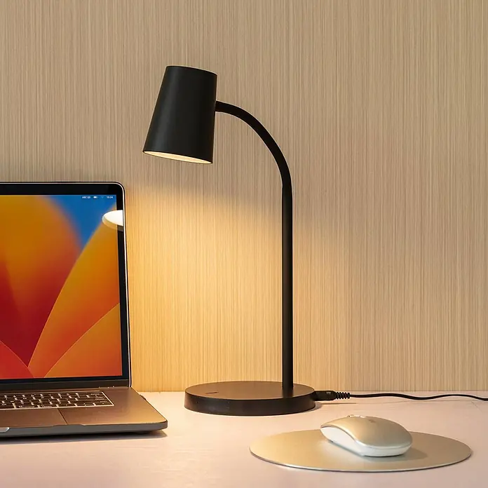Lindby Tischleuchte Ailina - schwarz, schwarz - Schwarze Schreibtischlampe mit gebogenem Stiel, die warmes Licht auf eine Holzwand und einen weißen Schreibtisch mit Laptop wirft.