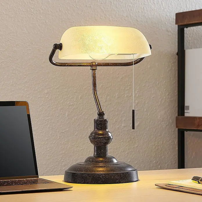 Lindby Tischleuchte Profina - L x B x H: 18,5 x 27,2 x 38,5 cm, braun / rost, creme / amber, E27 - Vintage-Bankerlampe in Altbronze-Optik mit beleuchtetem mattiertem Glasschirm auf einem Holzschreibtisch.