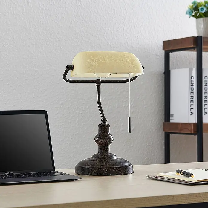 Lindby Tischleuchte Profina - L x B x H: 18,5 x 27,2 x 38,5 cm, braun / rost, creme / amber, E27 - Bankerlampe in Bronze-Optik mit cremfarbenem Glasschirm auf einem hellen Schreibtisch neben einem Laptop.