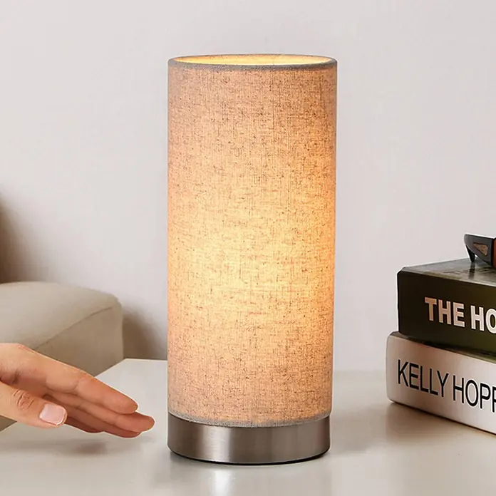 Lindby Tischleuchte Ronja - creme / amber, E14 - Eine zylindrische Tischlampe mit beigem Stoffschirm und gebürstetem Metallsockel, die warmes Licht abstrahlt. Eine Hand schwebt daneben.