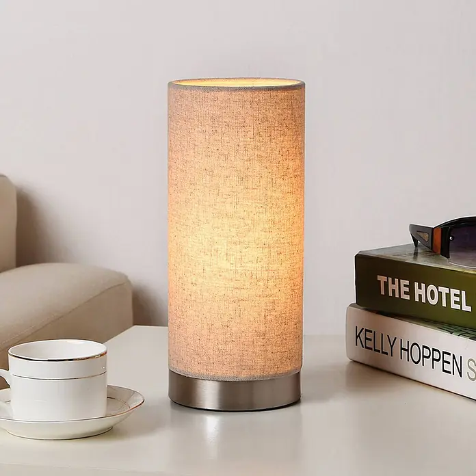 Lindby Tischleuchte Ronja - creme / amber, E14 - Zylindrische Tischlampe mit hellem Leinenlampenschirm und gebürstetem Metallsockel, leuchtend auf einem Beistelltisch.