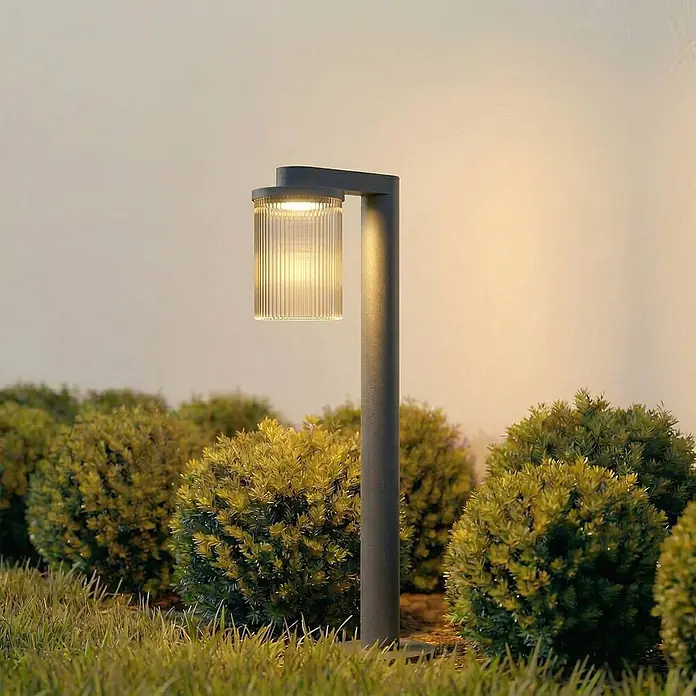 Lucande Außenlaterne Beelia -  - Moderne anthrazitfarbene Gartenlampe mit geripptem Glasschirm, strahlt warmes Licht aus, steht zwischen grünen Büschen in einer Gartenumgebung.