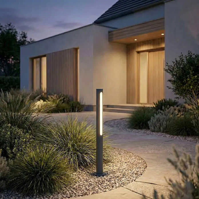 Lindby Außenlaterne Liravel - 8 x 8 x 60 cm - Dunkelgraue Pollerleuchte mit vertikalem LED-Streifen, die einen gepflasterten Gartenweg in der Dämmerung beleuchtet.