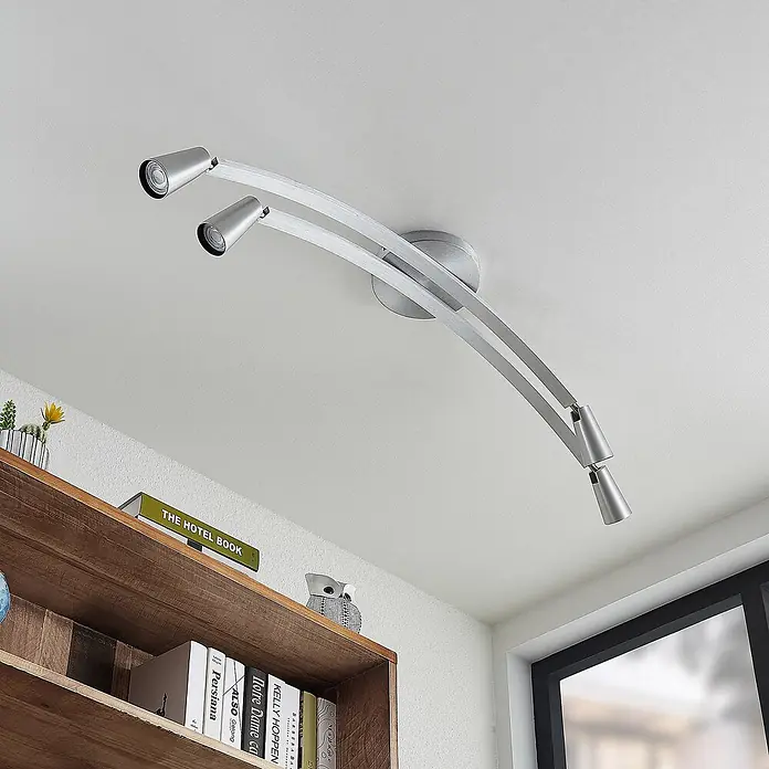 Lucande Deckenleuchte Velanoris - alu / grau / zink, alu / grau / zink, GU10 - Moderne silberne Deckenleuchte mit drei verstellbaren Strahlern an gebogener Schiene, beleuchtet einen Raum über einem Regal.