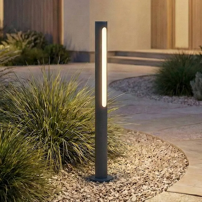 Lindby Außenlaterne Liravel - 8 x 8 x 60 cm - Moderne dunkelgraue Wegeleuchte mit vertikalem Leuchtstreifen, erhellt Gartenweg mit Kies und Pflanzen.