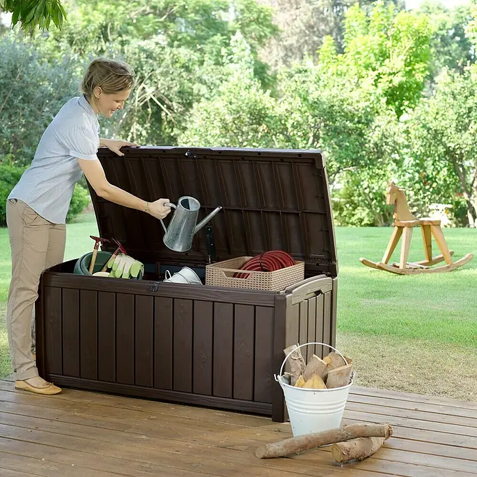 Keter Baúl para jardín Glenwood - Marrón, 65 x 128 x 61 cm, Polipropileno - Una mujer ordena una caja de jardín marrón en una terraza de madera, utilizada para guardar herramientas de jardinería.