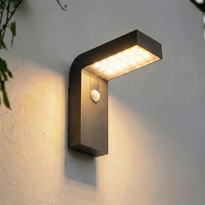 Prios Außenwandleuchte Aurino - 20,5 x 9 x 20 cm, schwarz, IP44 - Dunkelgraue Outdoor-LED-Wandleuchte mit Bewegungssensor, an einer strukturierten Wand montiert, leuchtend.