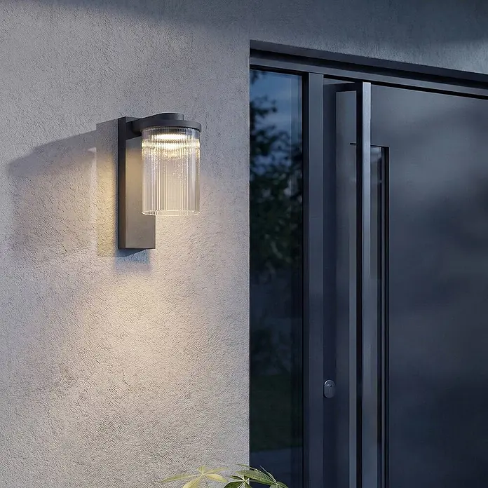 Lucande Außenwandleuchte Beelia - schwarz, klar / transparent, IP65 - Dunkelgraue Außenwandleuchte mit geripptem Glasschirm an strukturierter Wand neben moderner Tür, spendet warmes Licht.