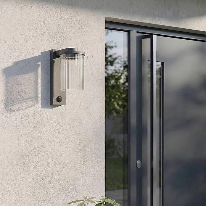 Lucande Außenwandleuchte Beelia - schwarz, klar / transparent, IP65 - Dunkelgraue Außenwandleuchte mit geripptem Glasschirm und Bewegungssensor, an einer Wand neben einer Tür montiert.