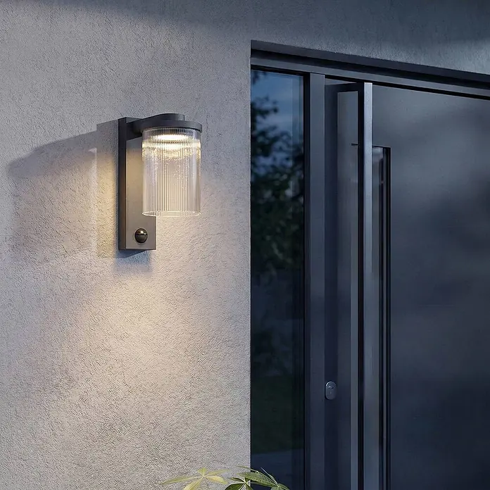 Lucande Außenwandleuchte Beelia - schwarz, klar / transparent, IP65 - Dunkelgraue Außenwandleuchte mit klarem, geripptem Glaszylinder und Sensor, beleuchtet eine strukturierte Wand neben einer dunklen Tür.