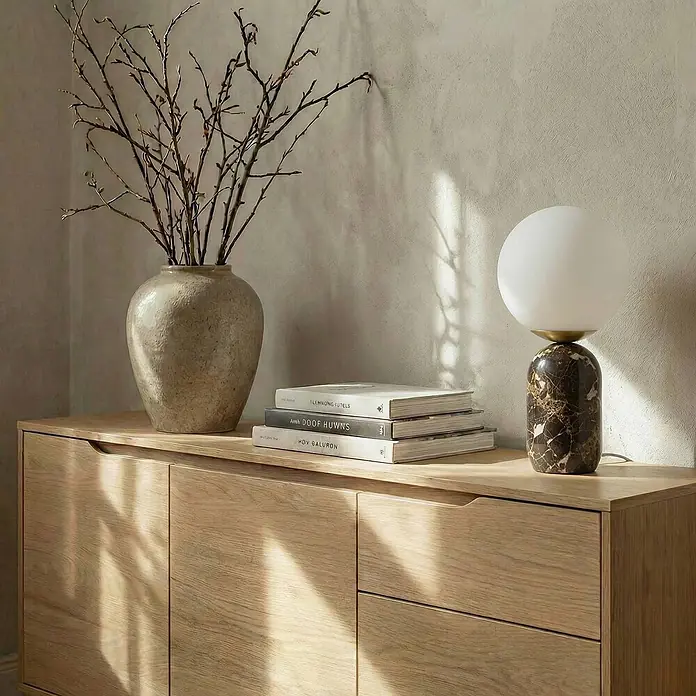 Lindby Tischleuchte Lennes - braun / rost, weiß / opal, E27 - Heller Holzschrank mit Vase voller Äste, gestapelten Büchern und einer Marmorlampe darauf, von Sonnenlicht beleuchtet.