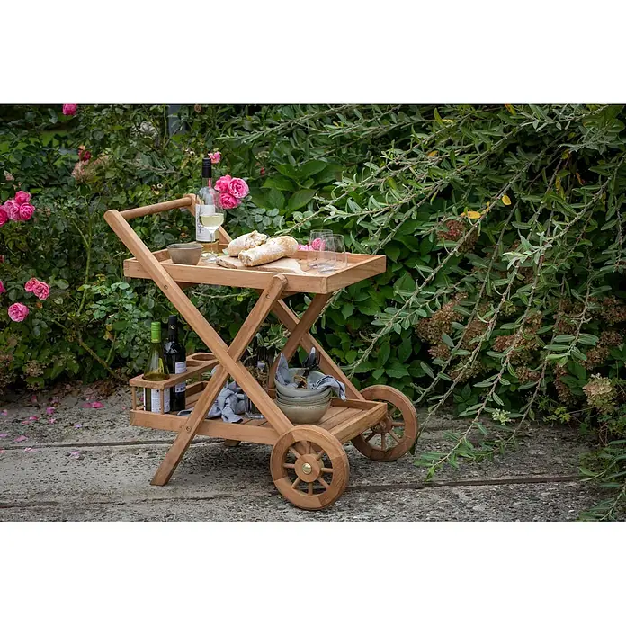 Bene Living Garten-Beistelltisch Chester - Teakholz, Rollbar, Rechteckig, Natur - Hölzerner Servierwagen mit zwei Ebenen und Rädern, beladen mit Wein, Brot und Geschirr in einem Garten.