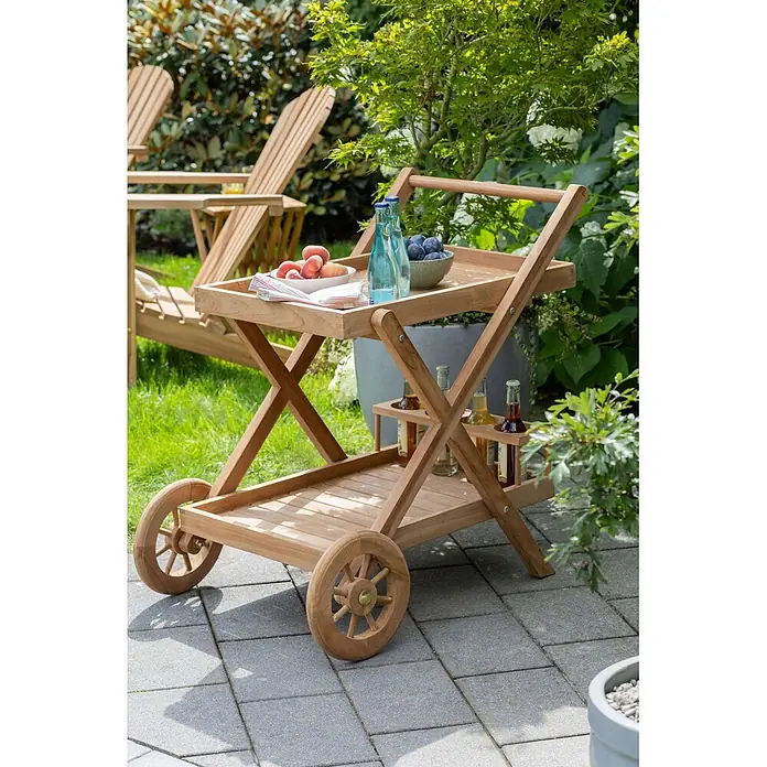 Bene Living Garten-Beistelltisch Chester - Teakholz, Rollbar, Rechteckig, Natur - Hölzerner Gartenwagen mit zwei Ablagen, beladen mit Obst und Flaschen, auf einer gepflasterten Terrasse im Garten.